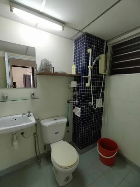 Bathroom features: white tiled walls, blue tiled accent wall, ceramic tile flooring, single-sink vanity with mirror, chrome basin mixer faucet, one-piece toilet, shower with handheld showerhead, wall-mounted shower faucet, shelf above sink, plastic bucket, window for ventilation, overhead fluorescent lighting.