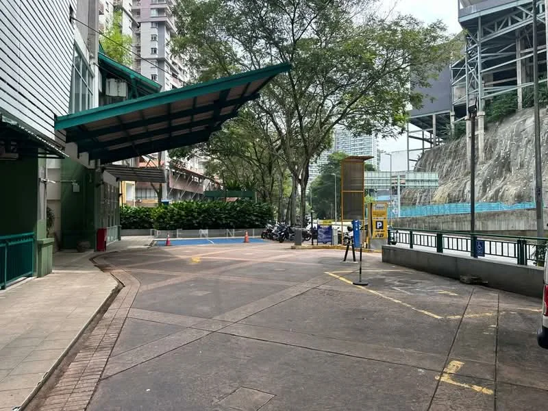 Exterior type: parking deck; features: covered parking area, concrete surface, green awning, landscaped greenery, security post, visible motorcycle parking, surrounding high-rise buildings, access road, perimeter fencing, blue construction netting; style: modern.