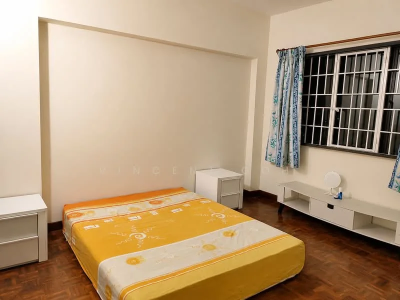 Bedroom features: queen-size bed with yellow bedspread, two white bedside tables, wooden flooring, window with white metal grille, blue patterned curtains, wall-mounted TV console, decorative mirror on console, plain white walls.