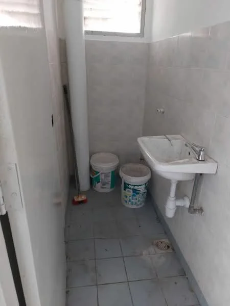 Bathroom features: white ceramic sink, chrome faucet, grey tiled walls, light grey floor tiles, drain in floor, two paint buckets, cleaning supplies, window for natural light.