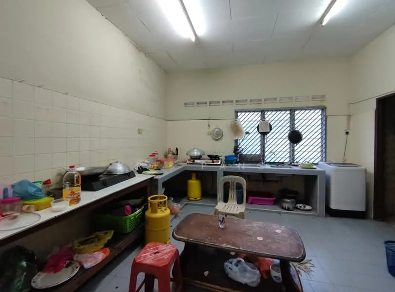 Kitchen features: white tiled walls, grey tiled flooring, stainless-steel sink, built-in countertop, kitchen cabinets, gas stove, exhaust hood, refrigerator, multiple cooking pots, plastic storage containers, dining table, plastic chairs, various utensils, window with metal grilles.