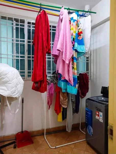 Laundry features: washing machine, clothes drying rack, various hanging clothes, tile flooring, window with metal grille, white walls, broom, shelf, light fixture.