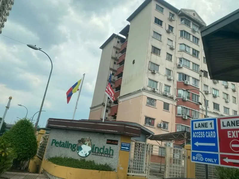 Exterior type: low-rise apartment block; features: cream painted facade, multiple air conditioning units visible, balconies on upper floors, entrance gate with signage, landscaped greenery at entrance, flags displayed at entrance, parking signage, security features visible; style: modern.