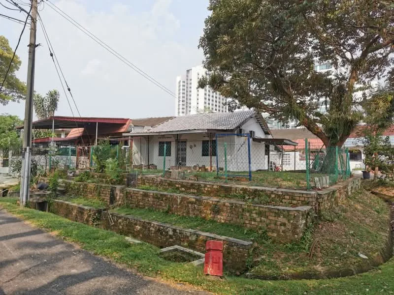 Exterior type: detached bungalow; features: single-storey building, pitched tile roof, white painted facade, brick wall foundation, green fencing around property, manicured lawn, mature trees, surrounded by greenery, driveway with interlocking pavers, adjacent to residential area, urban context with high-rise buildings in background; style: traditional.