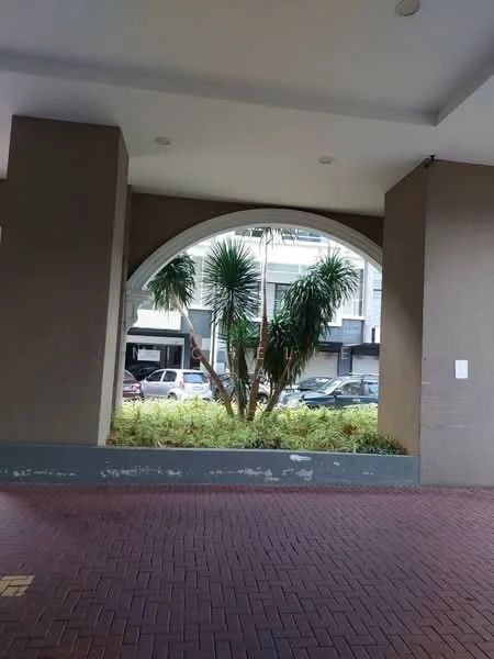 Exterior features: arched window opening, potted palm trees, landscaped area with greenery, brick pavement, concrete wall, overhang roof, visible cars in parking area; style: modern.