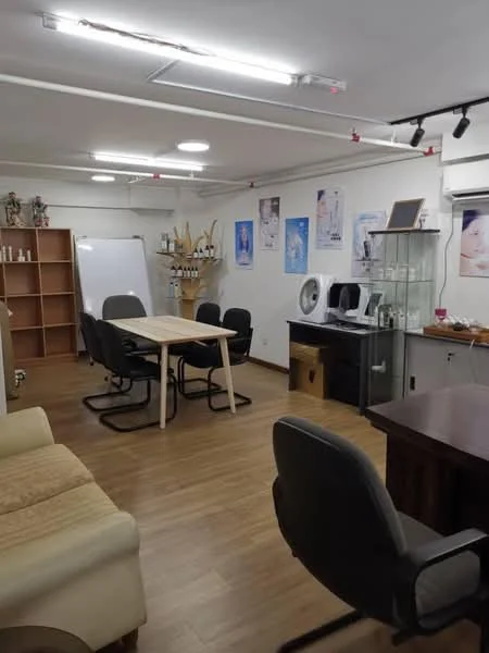 Room features: laminate flooring, white painted walls, recessed LED downlights, track lighting, air conditioning unit, glass display cabinet, wooden table, 4 black office chairs, sofa in cream color, shelving unit, whiteboard, decorative items on shelves; style: modern.