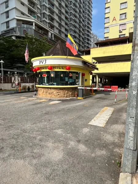 Exterior type: main entrance gate; features: yellow painted facade, stone cladding at base, roof with overhang, security booth, barrier gate, flagpole with flags, surrounding landscaping, paved driveway, visible parking area, surrounding high-rise buildings; style: modern.