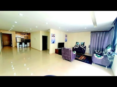 Living features: cream painted walls, glossy tile flooring, recessed downlights, floor-to-ceiling windows with sheer curtains, 2-seater fabric sofa in grey, 3-seater fabric sofa in grey, coffee table, TV console, wall-mounted TV, area rug with geometric pattern, decorative cushions, framed artwork; style: modern. Room features: open layout, kitchen area visible in background, dining table with chairs visible in background.