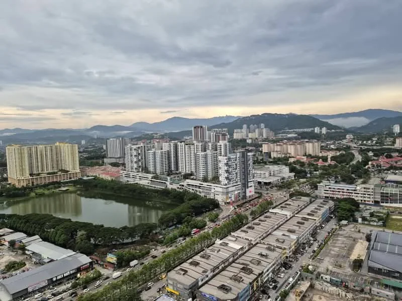 Exterior type: high-rise condominium; features: city skyline view, surrounded by greenery, near a lake, multiple high-rise buildings, commercial area at the base, mountain backdrop, urban landscape, cloudy sky; style: modern.