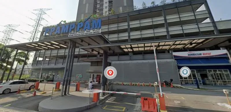 Exterior type: commercial building; features: multi-storey building, glass facade, entrance with awning, security boom gate, parking area, signage visible, surrounded by greenery, electricity pylons in background; style: modern.