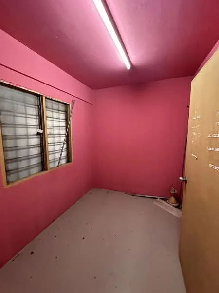 Room features: pink painted walls, plain ceiling, single window with grid pattern, wooden door, concrete flooring, fluorescent lighting.
