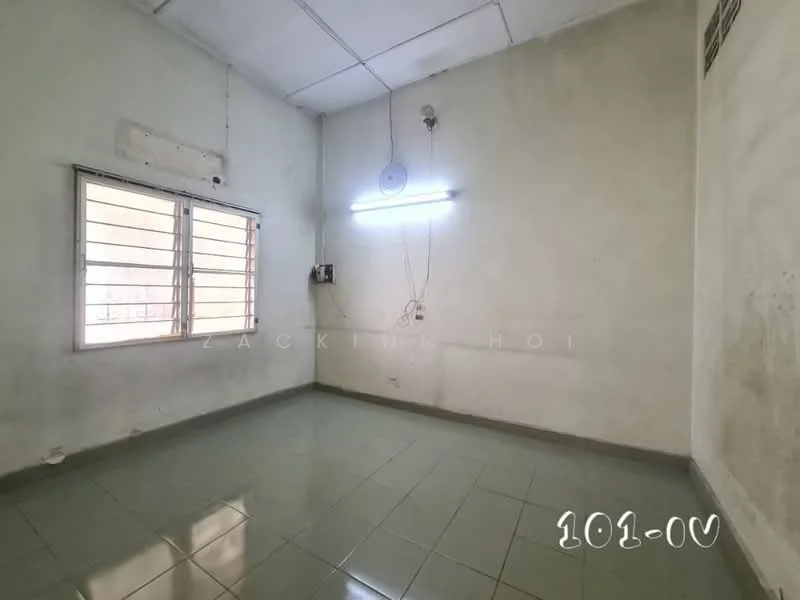 Room features: white painted walls, green ceramic tile flooring, single window with metal grille, fluorescent ceiling light, exposed electrical wiring, plain ceiling, no visible furniture or decor.