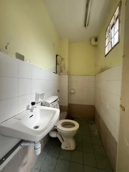 Bathroom features: white ceramic toilet, wall-mounted sink, chrome faucet, green tile flooring, white tiled walls, yellow painted walls, window with grid, exposed plumbing, fluorescent ceiling light.