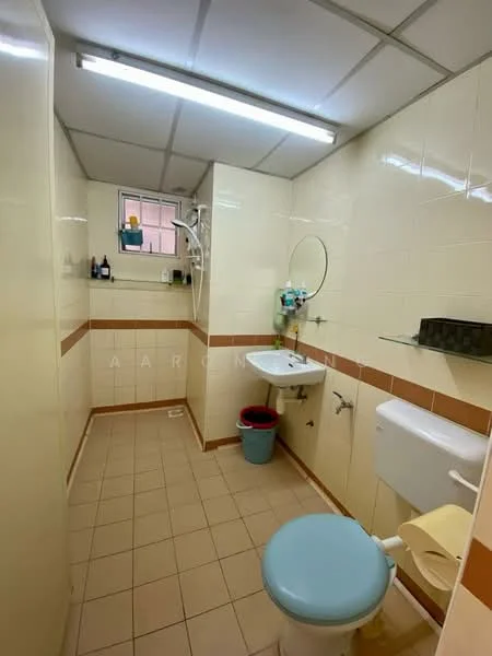 Bathroom features: white tiled walls, beige tiled accents, light beige floor tiles, single-sink vanity with mirror, chrome basin mixer faucet, toilet with blue seat, shower with wall-mounted showerhead, window for natural light, wall-mounted toiletries shelf, waste bin, toilet paper holder.