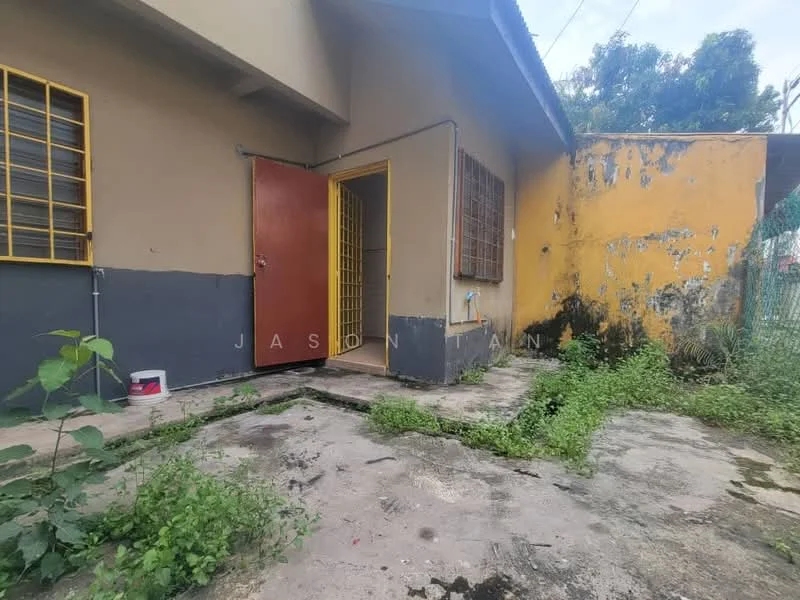 Exterior features: yellow painted wall, grey painted wall, metal door, window with metal grilles, overgrown vegetation, concrete flooring, fenced area, exposed utility piping.