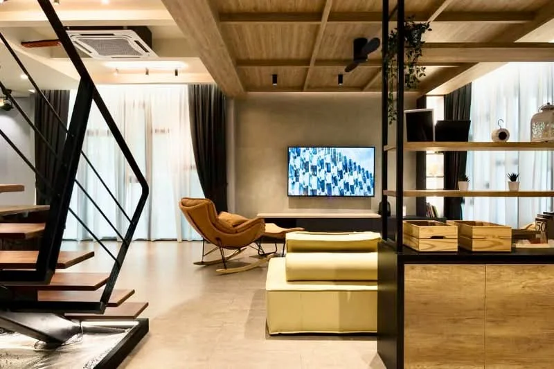 Living features: wooden ceiling with exposed beams, recessed downlights, floor-to-ceiling windows with sheer curtains, light grey painted walls, large wall-mounted TV, rocking chair in brown upholstery, yellow upholstered sofa, open shelving unit with decorative items, tile flooring in light beige, air conditioning unit mounted on ceiling, decorative plants, coffee table, area rug; style: modern.