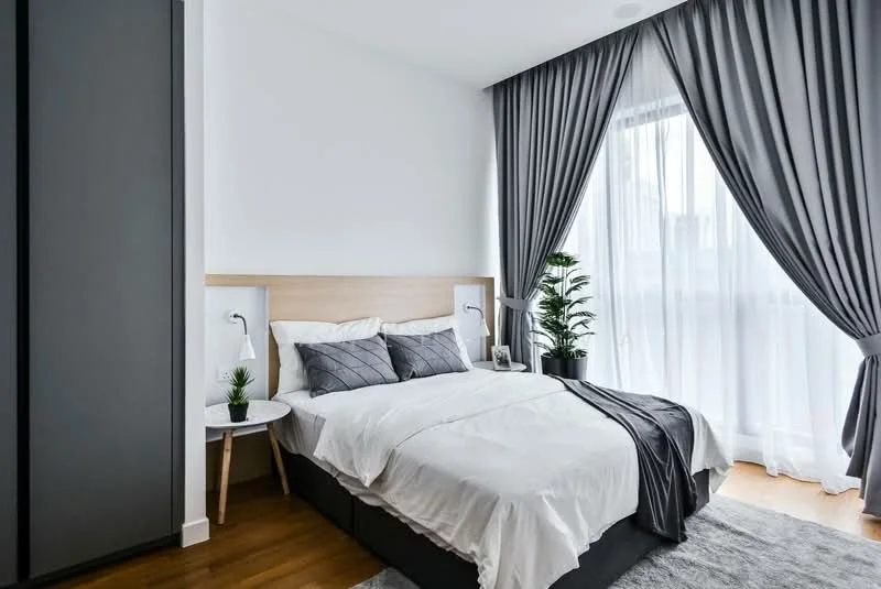 Bedroom features: queen-size bed with white bedding, upholstered headboard, nightstand with lamp, floor-to-ceiling windows, sheer curtains, blackout curtains, wooden flooring, decorative plant, area rug, wall-mounted reading lights, grey accent throw blanket; style: modern; view: city.