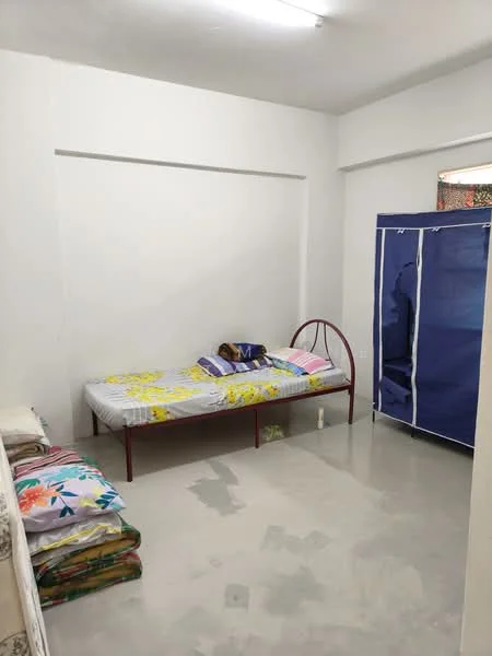 Bedroom features: single bed with floral bedding, white painted walls, concrete flooring, wardrobe with blue cover, window with natural light, decorative pillows on bed, folded bedding on floor, ceiling light fixture; style: minimalist.