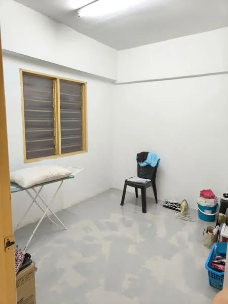 Room features: white painted walls, grey flooring, window with horizontal slats, ironing board, black plastic chair, blue laundry basket, plastic container, folded clothes on chair, ceiling light fixture.