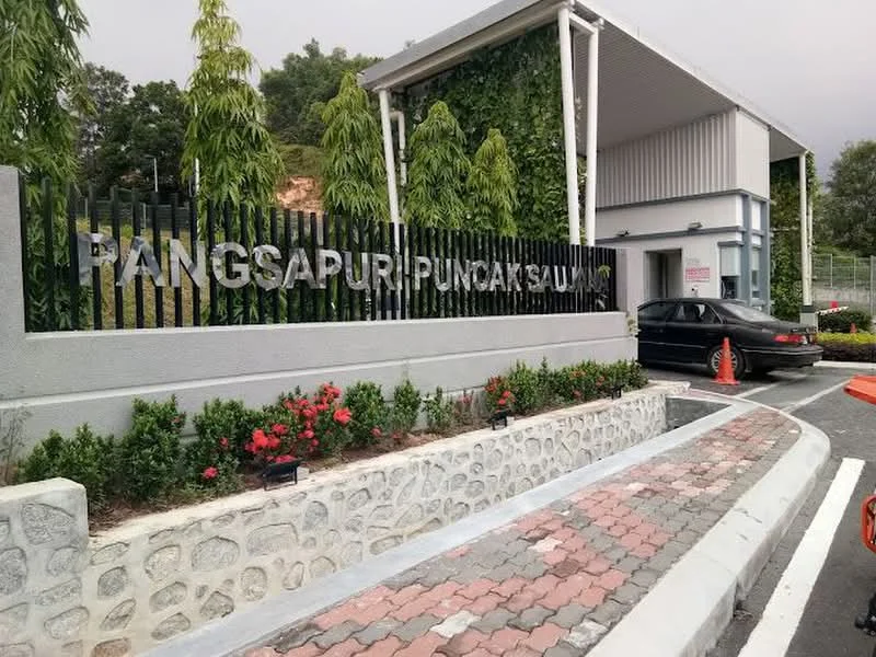 Exterior type: gated community; features: modern entrance gate, landscaped garden with shrubs, paved driveway, stone cladding on walls, signage with 'Pangsapuri Puncak Sau' in metal letters, surrounding greenery, parking area, decorative flower beds; style: modern.