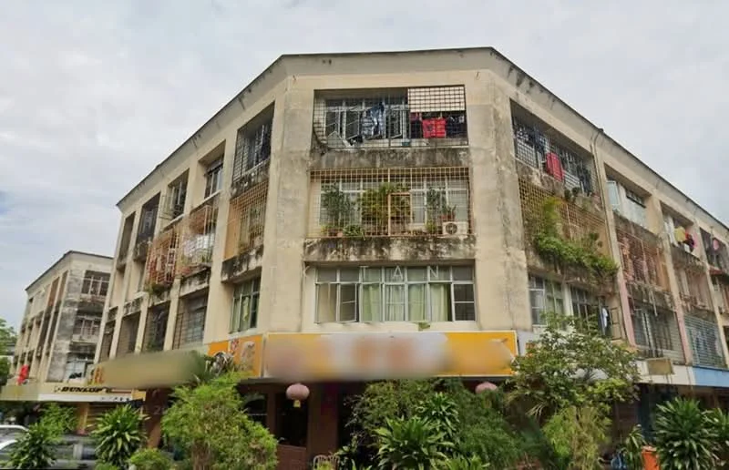 Exterior type: low-rise apartment block; features: weathered concrete facade, multiple balconies with metal grilles, visible air conditioning units, ground floor commercial space, overgrown plants at the base, laundry hanging on balconies, mature greenery surrounding the building, two-storey structure; style: traditional.