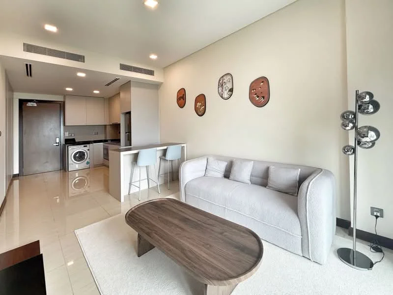 Living features: light grey fabric sofa, wood coffee table, area rug, wall-mounted TV, recessed downlights, decorative wall masks, open-plan layout with kitchen, tile flooring, floor-to-ceiling windows, white painted walls; style: modern. Kitchen features: white cabinetry, black countertop, built-in washing machine, bar seating with two stools, stainless-steel sink, chrome faucet, recessed lighting, ceramic tile backsplash; style: modern.