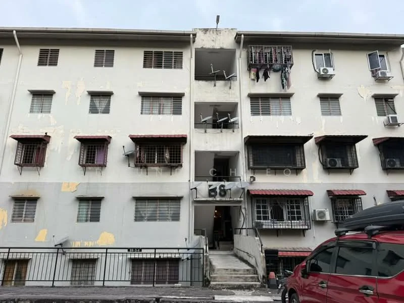 Exterior type: low-rise apartment block; features: white painted facade, multiple windows with metal grilles, red awnings over windows, visible air conditioning units, staircase leading to upper floors, balconies with laundry hanging, parking area in front, weathered paint, ground-level access; style: traditional.