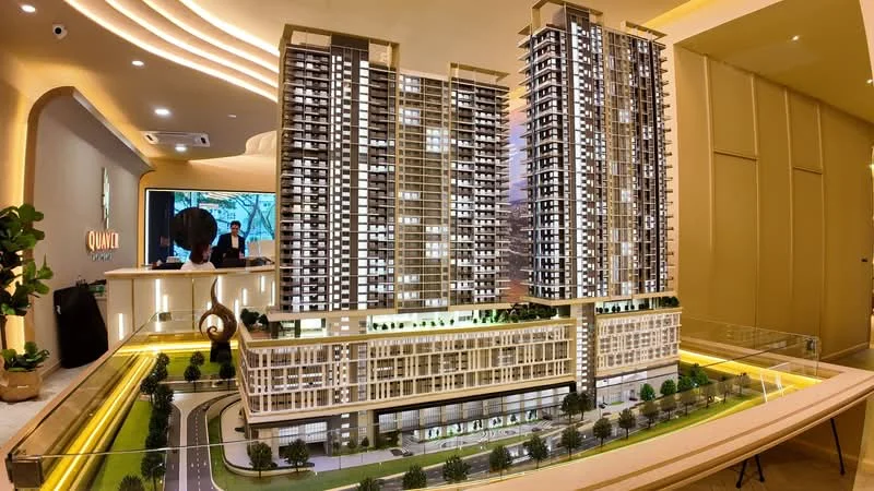 Exterior type: high-rise condominium; features: architectural model of building, multiple towers, landscaped podium, greenery on podium, roadway in front, illuminated base, modern design elements, glass facade, balconies on upper floors, recessed balconies, lobby area visible in background; style: modern.