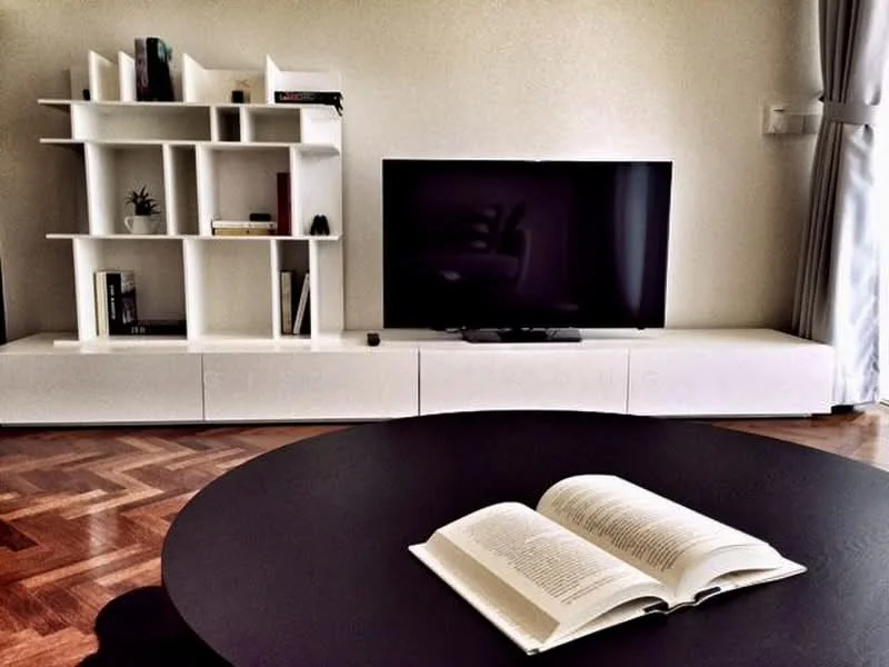 Living features: dark round coffee table, white TV console, black flat-screen TV, white bookshelf with open shelving, decorative plant on shelf, books on shelf, light-colored walls, herringbone wood flooring, curtains, natural light from window; style: modern.