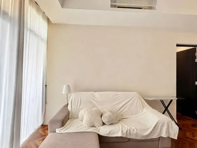 Living features: 3-seater fabric sofa in beige, white throw blanket, 2 decorative pillows, lampshade table lamp, wooden flooring, floor-to-ceiling windows with sheer curtains, exposed air conditioning unit, side table, dark wooden door; style: modern.
