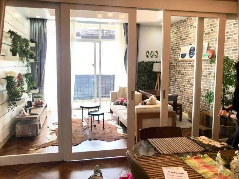 Living features: exposed brick wall, white painted walls, wooden flooring, sofa in cream, coffee table, area rug with animal print, shelves with plants, floor-to-ceiling windows, sliding glass door, curtains, decorative wall art, wall-mounted TV, greenery visible outside; style: modern; view: garden.
