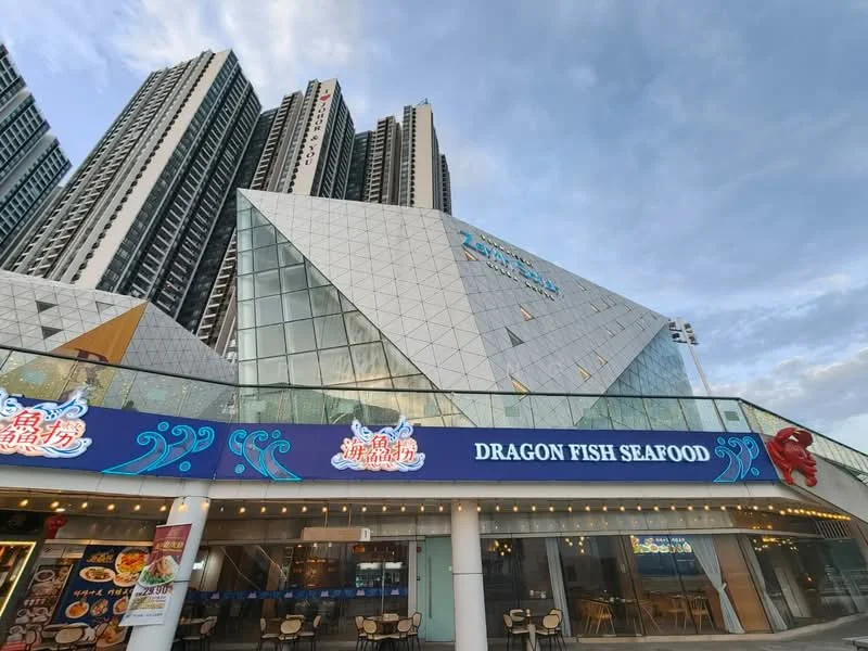 Exterior type: commercial building; features: glass facade, triangular architectural design, outdoor dining area, signage for Dragon Fish Seafood, multiple storeys, surrounded by high-rise buildings, modern design elements; style: contemporary.