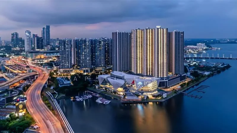 Exterior type: high-rise condominium; features: glass curtain wall, illuminated facade, surrounded by water, adjacent to highway, city skyline in background, multiple high-rise buildings, marina with boats, urban landscape, night view, manicured waterfront, roadway with traffic; style: modern.