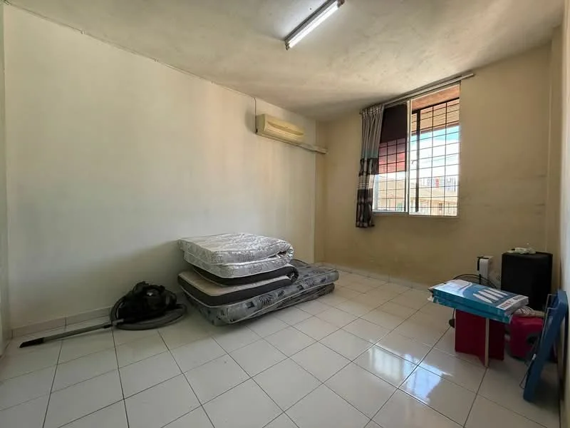 Room features: white painted walls, tile flooring, window with grid pattern, air conditioning unit, two stacked mattresses, vacuum cleaner, small table, plastic chair.