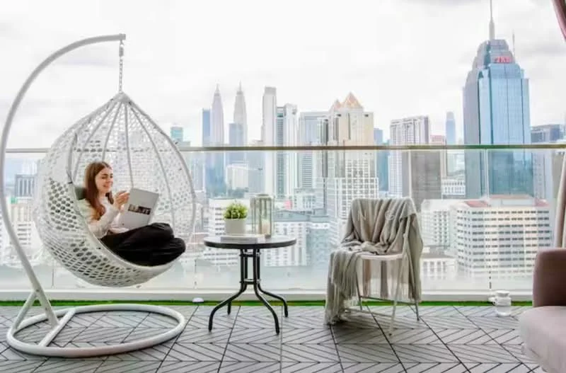 Balcony features: hanging rattan egg chair, small round table, single chair, city skyline view, glass railing, outdoor flooring, decorative plant, throw blanket; style: modern; view: city.