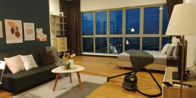 Living features: dark grey accent wall, light wood flooring, floor-to-ceiling windows, curtains in dark brown, 3-seater fabric sofa in black, round coffee table with white top, area rug in light grey, decorative wall art, TV console, vacuum cleaner, bed with grey bedding; style: modern; view: city.