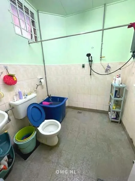 Bathroom features: light green painted walls, cream tiled walls, grey tiled flooring, toilet, shower with handheld showerhead, plastic storage shelf, blue plastic bucket, green plastic bucket, wall-mounted mirror, wall-mounted soap dispenser, various toiletries on shelf.
