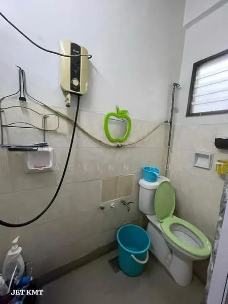 Bathroom features: white tiled walls, light grey floor tiles, one-piece toilet, green toilet seat, shower head with wall-mounted holder, water heater, small window for ventilation, plastic bucket, wall-mounted shelf, green shower curtain, soap dispenser.