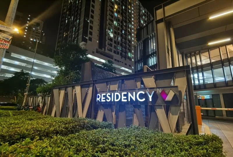 Exterior type: high-rise condominium; features: illuminated building sign, modern facade with geometric design, landscaped greenery in front, surrounding high-rise buildings, nighttime lighting, glass and metal elements, parking structure visible in background; style: modern.
