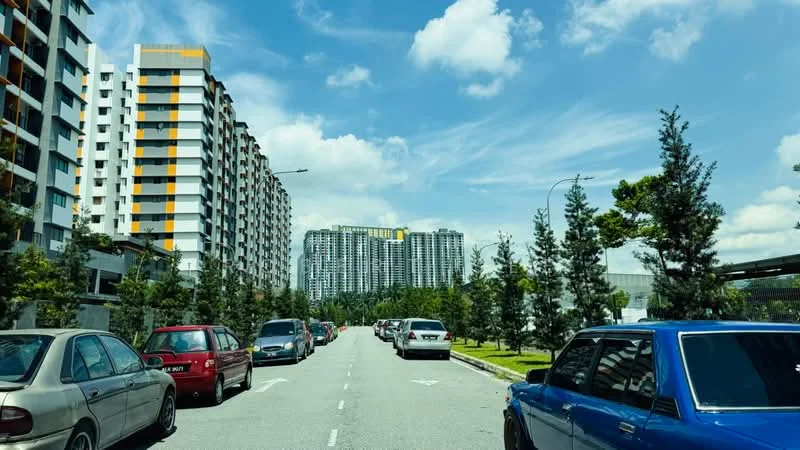Exterior type: high-rise condominium; features: multiple high-rise buildings, landscaped greenery along the road, paved road, parking spaces visible, trees lining the street, clear blue sky, urban setting; style: modern.