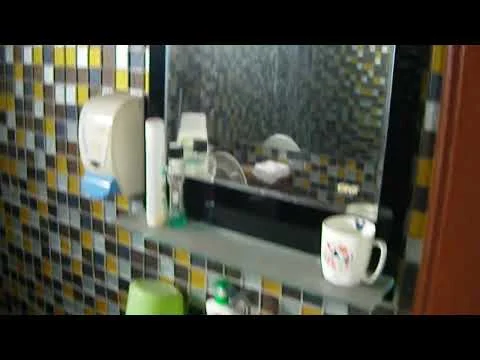 Bathroom features: mirror, shelf, soap dispenser, toothbrush holder, mug, various toiletries, tiled walls with colorful mosaic pattern.