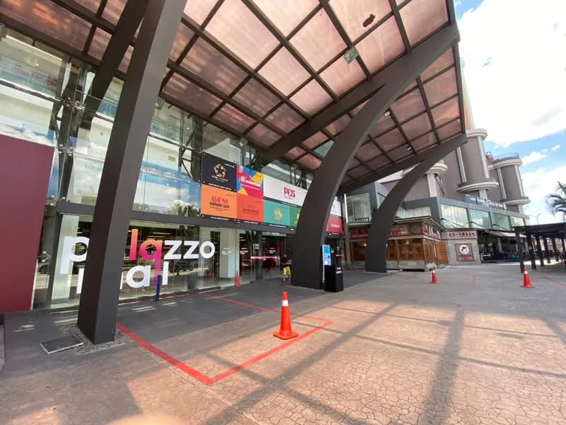 Exterior type: shopping mall; features: glass facade, metal awning, large entrance signage, paved walkway, traffic cones, outdoor seating area, surrounding commercial units, visible storefronts, canopy structure, modern architectural design; style: modern.
