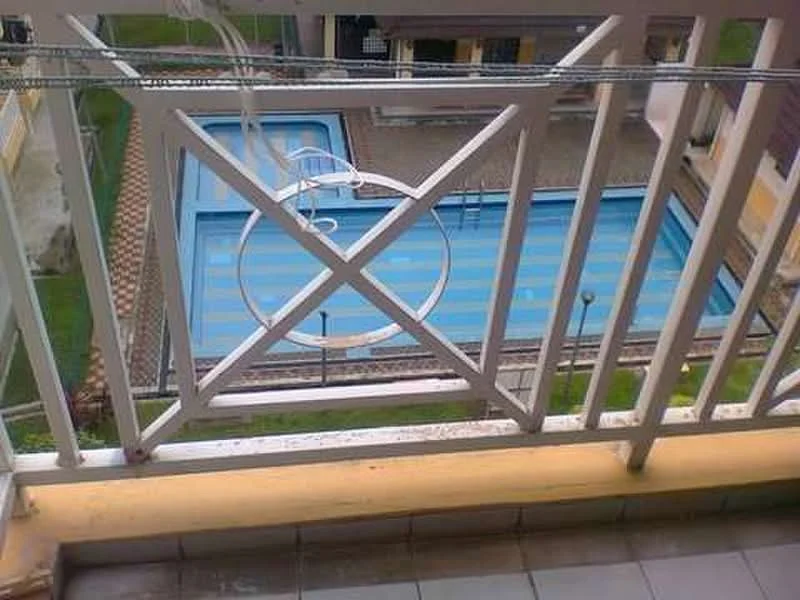 Balcony features: white railing, grey tiled flooring, view of swimming pool, green lawn, surrounding buildings; view: pool.