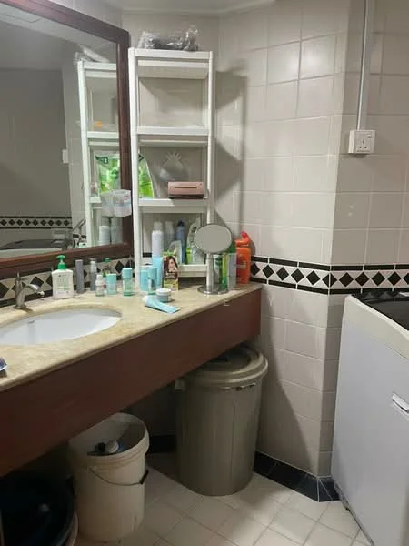 Bathroom features: single-sink vanity with beige countertop, wood cabinet under sink, mirror above sink, white ceramic sink, chrome basin mixer faucet, black and white tiled backsplash, white tiled walls, light-colored tiled flooring, built-in shelving unit, various toiletries and personal care items on shelves, waste bin, washing machine.