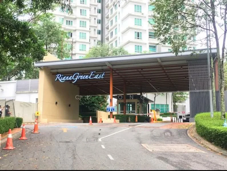 Exterior type: gated community; features: covered entrance with wooden beams, signage with 'Riana Green East', manicured landscaping, paved driveway, security checkpoint, traffic cones, surrounding greenery, multi-storey residential building in background; style: modern.