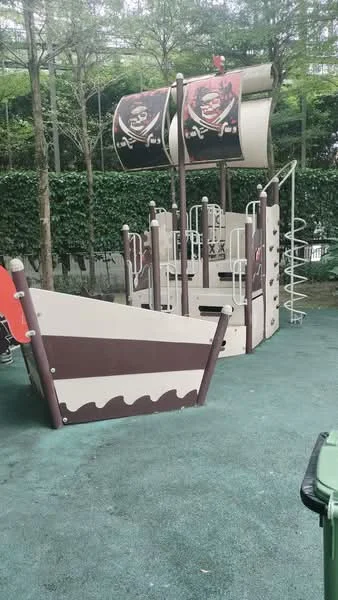 Exterior type: children's playground; features: pirate ship-themed play structure, two sails with pirate flags, slide, climbing structure, safety railings, soft rubber flooring, surrounding greenery, fenced area; style: modern.