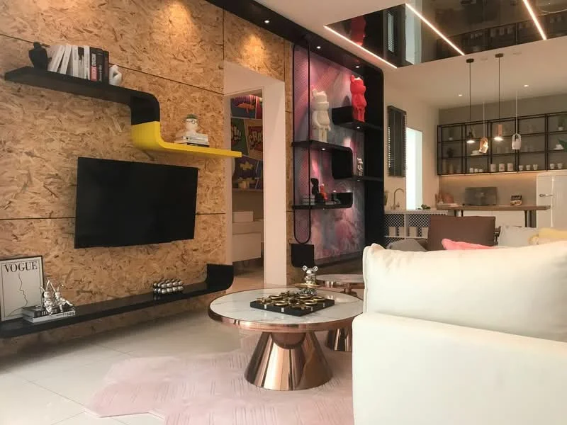Living features: white leather sofa, round coffee table with rose gold base, black wall-mounted TV unit, yellow accent shelf, wood panel accent wall, decorative figurines on shelves, framed artwork on wall, recessed LED lighting, pink area rug, open-plan layout to kitchen, glass display shelves, pendant lighting above kitchen area; style: modern.