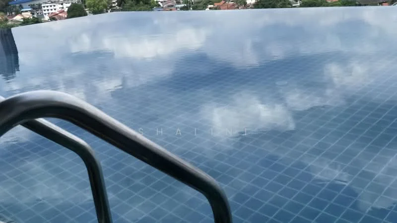 Exterior type: swimming pool deck; features: infinity pool, tile flooring around pool, metal railing, clear blue water, reflection of clouds on water, surrounding greenery, urban backdrop; style: modern.