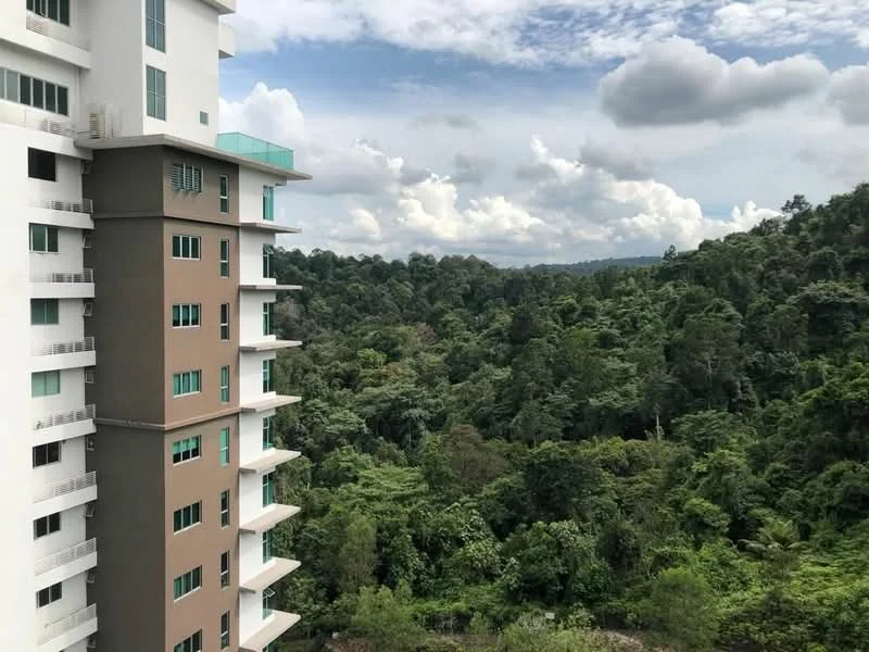 Exterior type: high-rise condominium; features: glass balcony railings, cream painted facade, brown accent panels, multiple floors visible, surrounded by greenery, urban view, cloudy sky, lush forest in background; style: modern.