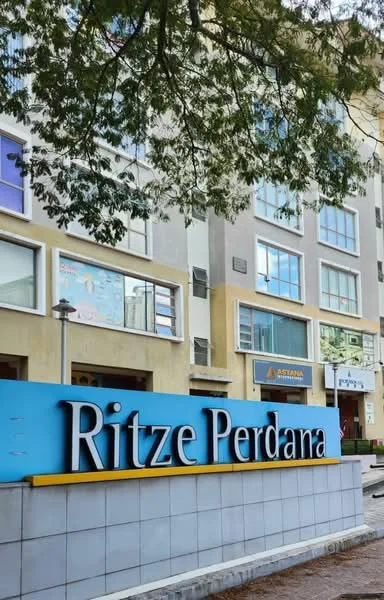 Exterior type: low-rise apartment block; features: blue signage with 'Ritze Perdana', multiple windows, cream painted facade, ground-level landscaping, street view, trees in foreground; style: modern.